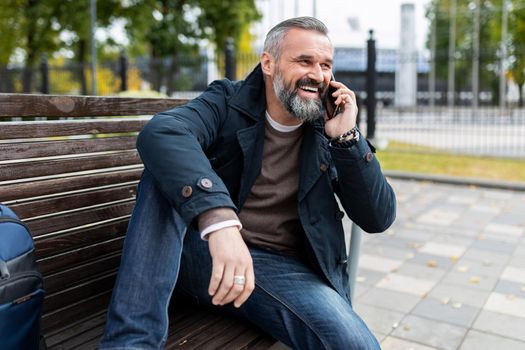 a mature man with a gray beard is talking on the phone with a smile while sitting on a bench in a city park