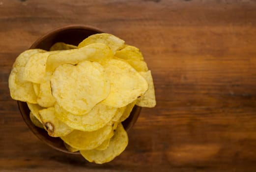 Potato chips in bowl on a wooden background, top view. Salty crisps scattered on a table.