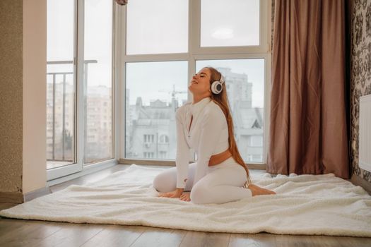 Side view portrait of relaxed woman listening to music with headphones lying on carpet at home. She is dressed in a white tracksuit.