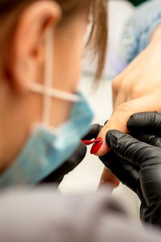Manicure varnish painting. Close-up of a manicure master wearing rubber black gloves applying red varnish on a female fingernail in the beauty salon.