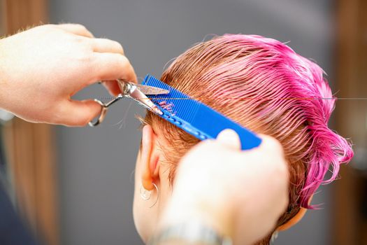 Professional hairstylist is cutting short pink hair with scissors in hair salon close up.