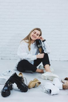 A young woman sits on the floor among shoes and chooses new shoes for herself. Lady in a white shirt and black trousers. Woman in white shoes.