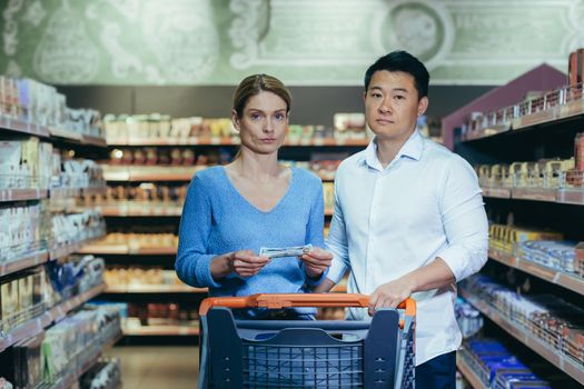 Upset young couple in supermarket, man and woman looking at camera poor have no opportunity and money to shop, multiracial family