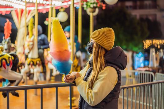 Young woman holding a cotton candy and smiling at a Christmas fair wearing a yellow wool cap wears a medical mask against coronavirus COVID 19