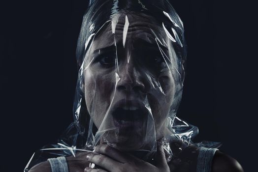 Fighting for oxygen, fighting for her life. A young woman suffocating with her head wrapped in plastic while isolated on a black background.
