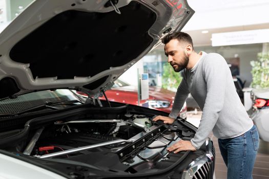 Attentive buyer in a car dealership examines the engine compartment and engine characteristics of a car