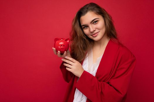 Young beautiful european stylish brunette woman wearing white blouse and red cardigan isolated over red background with positive sincere emotions. Holding little red moneybox. Free space