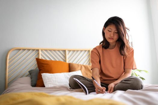 Pensive young Asian woman sitting on bed with serious expression writing on journal in cozy bedroom. Copy space.