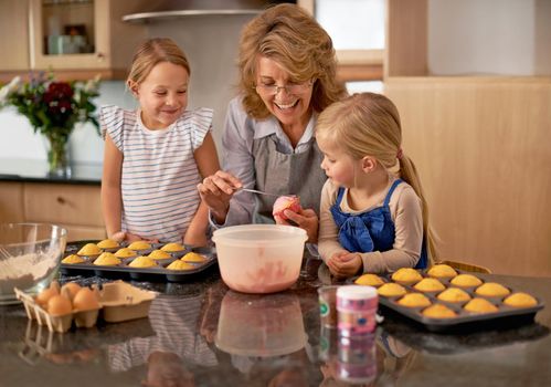 Teaching her grandchildren valuable kitchen skills. Two little girls baking cupcakes with the help of their grandmother at home.