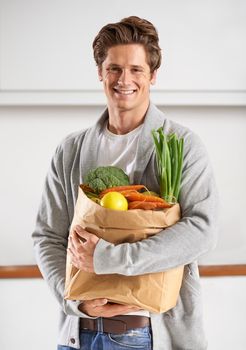 Only organic for me. A young man in a kitchen holding a brown paper bag filled with vegetables.