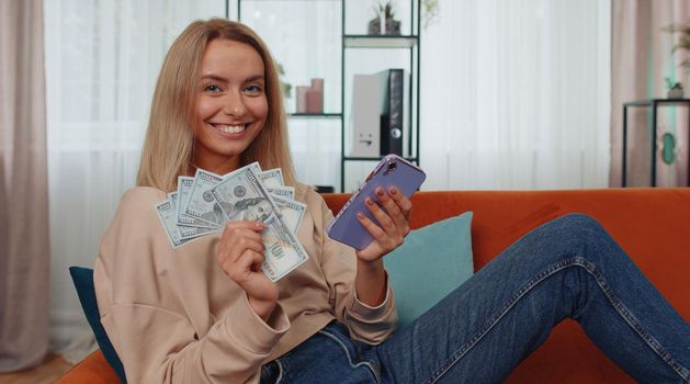 Smiling happy young woman at home counting money cash use smartphone, income, saves, lottery win