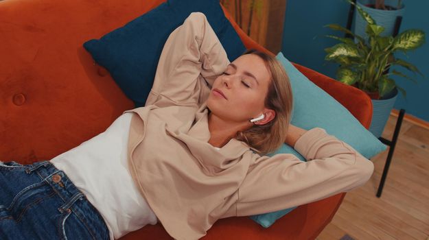 Happy young woman in wireless earphones relaxing lying on sofa at home listening favorite music