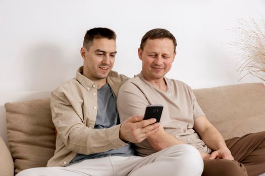 Happy gay couple with casual clothes holding smartphone and talking with friends online, having video call. Homosexual relationships and alternative love.