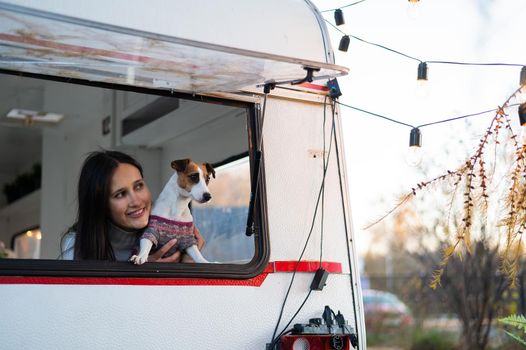 Caucasian woman looks out of the window of the van while hugging her dog Jack Russell Terrier. Travel in a camper in autumn.