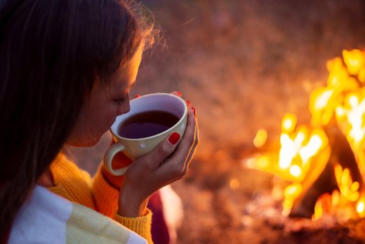 Girl drinking warm tea sitting near bright bonfire