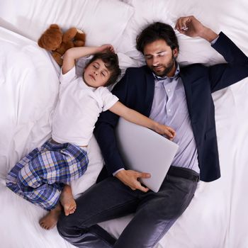 Being a single father can be exhausting. A father and son sleeping on a bed while his father is still fully dressed and holding a laptop.