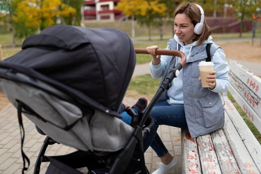 mother with a baby in a stroller sits on a bench and drinks coffee in headphones