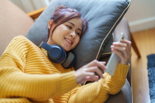 Portrait of a smiling Asian woman wearing a pair of headphones and using her phone and listening to music while sitting on the sofa