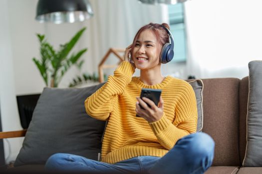 portrait of a young Asian woman woman using headphones holding smartphone while sitting on sofa