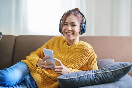 Portrait of a smiling Asian woman wearing a pair of headphones and using her phone and listening to music while sitting on the sofa
