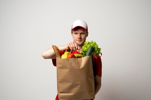 Food shipping, profession and people concept. Delivery man in red uniforn holding a paper bag full of vegetables.