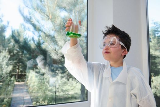 Adorable dark-haired child boy wearing safety goggles and lab coat examining the chemical reaction going on in the flask