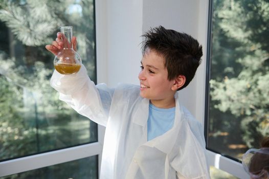 Happy mischievous smart schoolboy in white lab coat, examining the chemical reaction going on in the flat bottomed flask