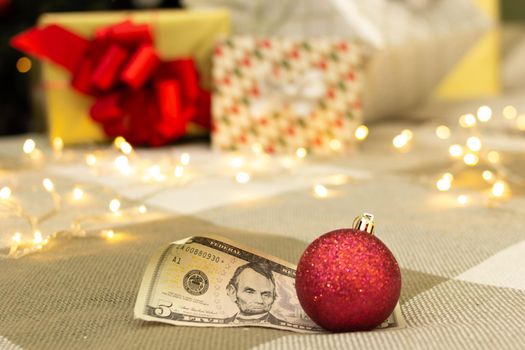 Dollar bill with red christmas decoration, gifts with lights and xmas tree on blurred background, festive greeting card