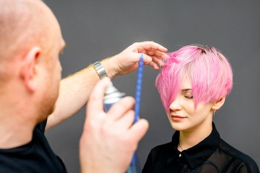 The hairdresser is using hair spray to fix the short pink hairstyle of the young caucasian woman in the hair salon.