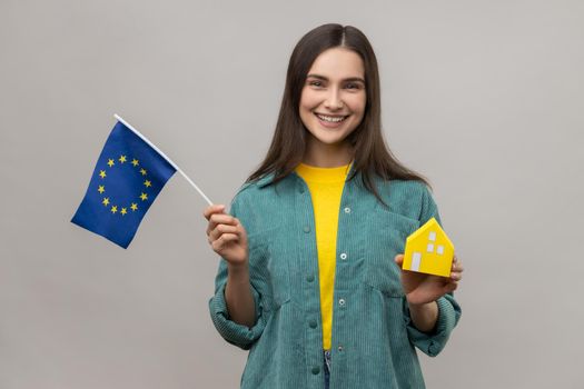 Satisfied holding european flag and paper house, dreaming to buy accommodation in Europe.