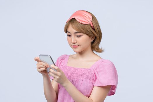 Happy young woman wears sleepmask uses mobile phone for sending text messages over gray background