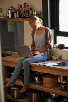 Recipe blogging. a beautiful woman sitting on her kitchen counter using a laptop.