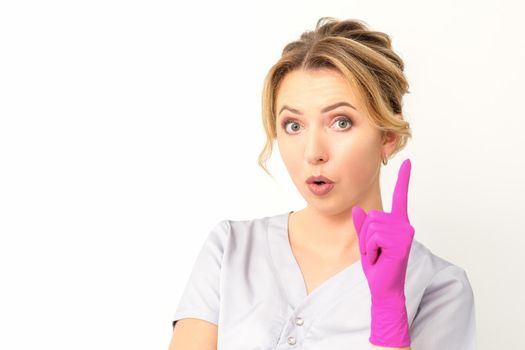 Young caucasian woman doctor wearing pink gloves keeping index finger pointed upwards making a gesture with index finger. I have an idea.