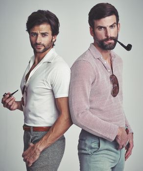 Theyve got far out style. Studio shot of two men standing together while wearing retro 70s wear and smoking pipes.