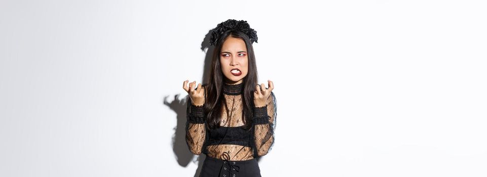 Image of annoyed and bothered asian woman looking mad, losing temper, clenching fists mad and rolling eyes, standing in evil witch costume on halloween, white background