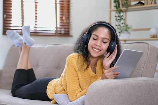 african young woman relaxing at home lying on sofa and listening to music on tablet wearing headphones. girl relaxing on the sofa of a cozy home