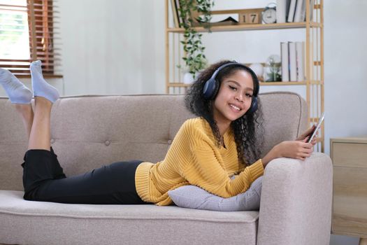 african young woman relaxing at home lying on sofa and listening to music on tablet wearing headphones. girl relaxing on the sofa of a cozy home