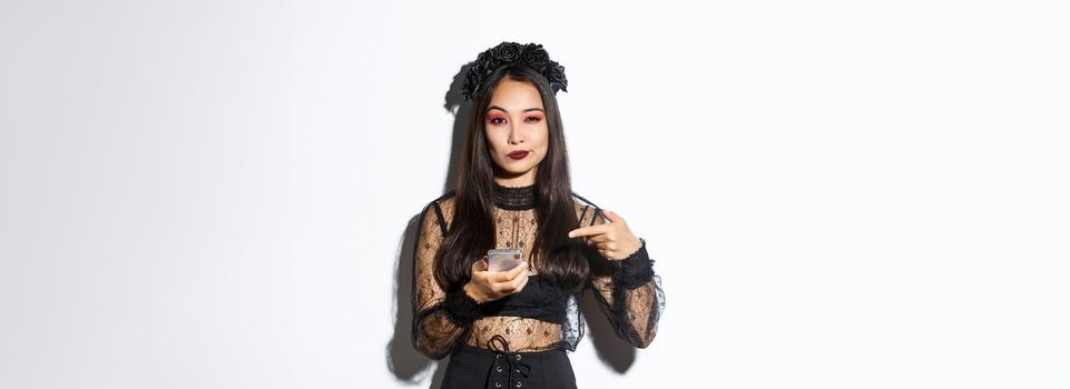 Sassy beautiful asian woman in gothic dress and black wreath pointing finger at mobile phone, showing something about halloween