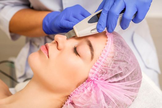 Young caucasian woman receiving facial skin cleaning by ultrasonic cosmetology face equipment in a medical salon.