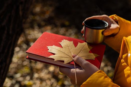 Woman holding thermos of hot tea red book and yellow fallen maple leaf in autumn.