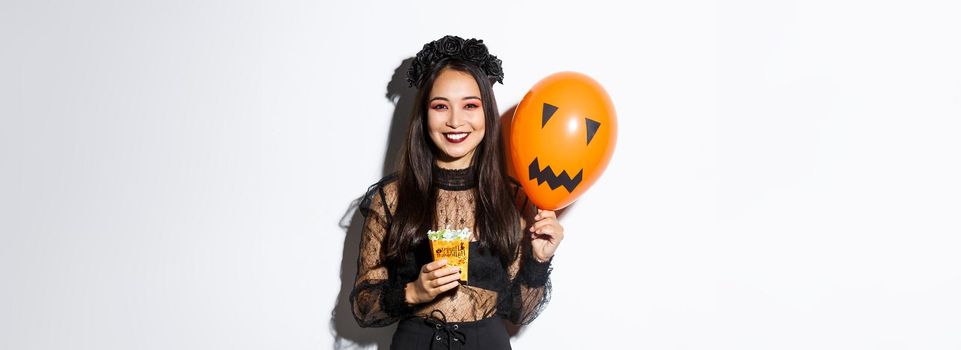 Image of cheerful asian girl trick or treating on halloween, holding sweets and orange balloon with scary face