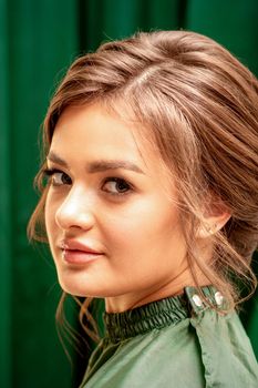 The fashionable young woman. Portrait of the beautiful female model with long hair and makeup with closed eyes. Beauty young woman with a brown curly hairstyle on the background of a green curtain