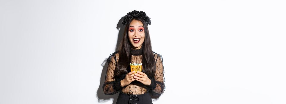Beautiful asian girl smiling happy, holding sweets, wearing witch costume on halloween, enjoying trick or treating