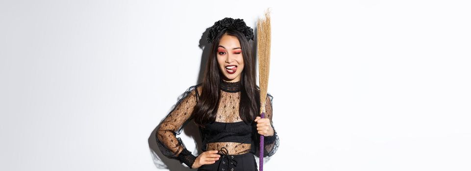 Sassy good-looking asian woman in witch costume showing tongue, holding broom and posing over white background