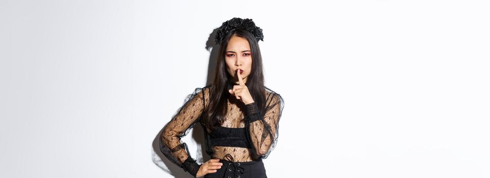 Mysterious asian witch shushing at you with concerned face, hiding secret, tell be quiet. Woman in undead widow costume prepare surprise for halloween, standing over white background