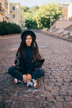 Young woman in hat sitting at city on cobblestone
