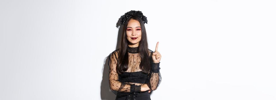 Creative young woman in witch costume smiling pleased as have great idea, raising finger to say suggestion. Female asian dressed-up like widow or mysterious magician, white background