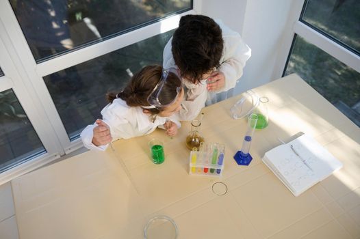 Overhead view of school kids in safety goggles and white lab coat, learning Chemistry in the school science laboratory