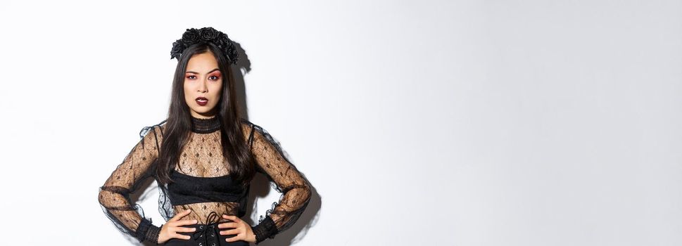 Angry and pissed-off asian female magician, evil witch in black dress and wreath looking mad at someone, frowning and looking judgemental, standing over white background