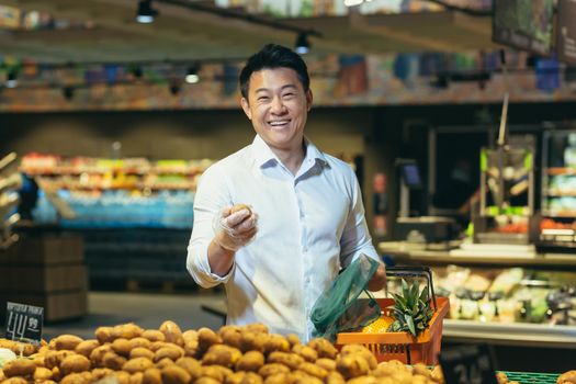 An Asian man is standing in a supermarket in the vegetable section, holding potatoes in his hands